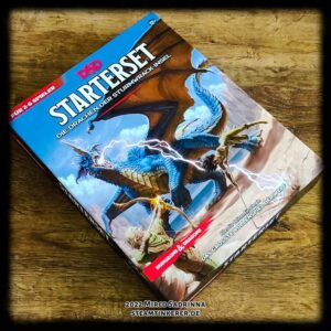 Die Box des D&D Startersets "Die Drachen der Sturmwrack-Insel". Ein riesiger blauer Drache ist im Kampf gegen zwei Held*innen verwickelt und speit Blitze aus seinem Maul.