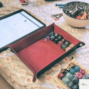 Blick auf den Spieltisch. Darauf befindet sich ein Tablet sowie eine Würfelunterlage. In diesem sowie daneben befinden sich mehrere Würfel. Außerdem ist eine Schüssel mit Süßigkeiten zu sehen.