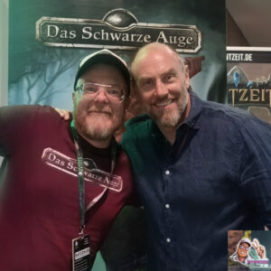 Ich stehe zusammen mit dem Niko Hoch, DSA-Chefredakteur, vor einem DSA-Aufsteller. Ich selbst trage ein T-Shirt mit der Aufschrift "Das Schwarze Auge".